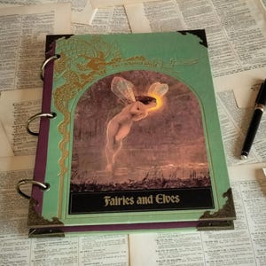May include: A journal titled "Fairies and Elves" with a green cover and a picture of a fairy. The journal has metal corner protectors and rings. A pen rests on a page of a dictionary.