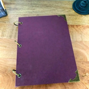 May include: A purple journal with brass-colored corner protectors and three ring binder closures. The journal is open, displaying the blank pages. The journal is on a wooden surface.