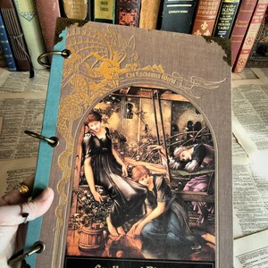 May include: A vintage-style journal with a brown cover and gold accents, featuring an illustration of women in flowing gowns. The journal is titled "The Enchanted World" and "Spells and Bindings". The journal is held open with brass rings, and is surrounded by antique books.