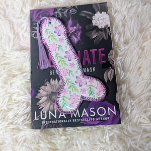 May include: A floral bookmark shaped like a phallus, with pink and purple flowers and green leaves. It has a purple tassel and is placed on a book. The book's title is "Ultimate Mask" by Luna Mason. The bookmark is handmade.