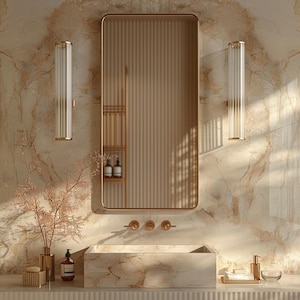 May include: A bathroom interior featuring a rectangular gold-framed mirror, a marble sink, and two glass and gold sconces. The wall is a light beige with a marble pattern. Decorative items include a vase with branches and bottles.