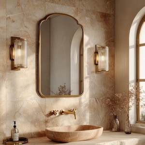 Puede incluir: Un área de lavabo de baño con un lavabo de piedra beige, un grifo de pared de latón con dos manijas en cruz y un espejo arqueado con un marco fino de latón. Dos apliques de pared cilíndricos de vidrio estriado flanquean el espejo sobre el lavabo.