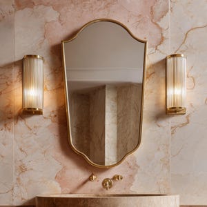 Peut inclure: Intérieur de salle de bain avec un miroir doré et un lavabo décoratif. Deux appliques murales avec du verre cannelé et des détails dorés encadrent le miroir. Le mur présente un motif de marbre rose et beige.