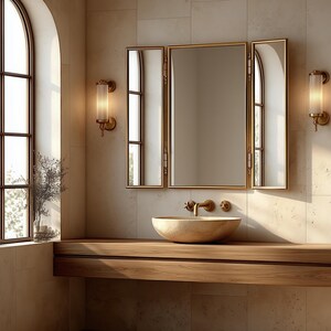 Peut inclure: Un miroir triptyque avec un cadre doré fin monté au-dessus d'une vasque en pierre. La vasque repose sur un meuble-lavabo en bois suspendu. Deux appliques murales cylindriques à abat-jour en verre strié encadrent le miroir, diffusant une lumière chaude.