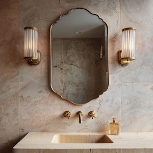 Puede incluir: Interior de baño con espejo dorado de borde festoneado sobre lavabo de piedra. Dos apliques de pared con pantallas de vidrio acanalado flanquean el espejo. Grifería dorada y dispensador de jabón.