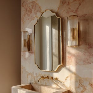 Peut inclure: Intérieur de salle de bain avec un miroir doré et des appliques murales assorties avec des abat-jours en verre cannelé. Le lavabo et le robinet sont dorés, sur un mur à effet marbre. Un verre est posé sur le bord du lavabo.