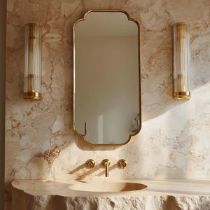 May include: A bathroom vanity with a gold-framed mirror, gold sconces, and gold faucet fixtures. The vanity has a stone countertop with a built-in sink. The wall is a light beige marble.