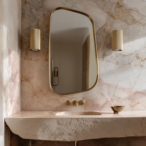 Op de afbeelding: Een moderne badkamer met een grote, onregelmatig gevormde, goudomrande spiegel. Twee gouden wandkandelaars flankeren de spiegel, en een stenen wastafel met gouden kranen staat eronder. De muren zijn bedekt met een roze en wit marmerpatroon.