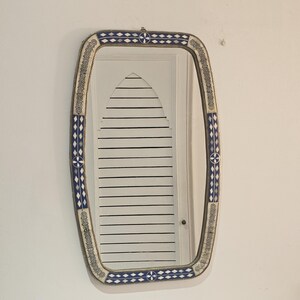 Peut inclure: Miroir mural rectangulaire avec un cadre décoratif. Le cadre présente un motif de formes géométriques bleues et blanches, avec une bordure dorée. Le miroir est accroché à un mur blanc.