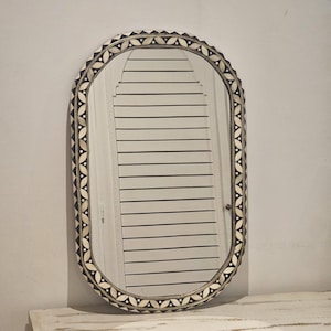 Peut inclure: Miroir ovale avec un cadre décoratif orné d'un motif de formes en forme de feuilles noires et blanches. Le miroir est monté sur une armoire blanche à deux portes et des boutons en laiton. Le miroir reflète une porte blanche avec des lignes horizontales noires.