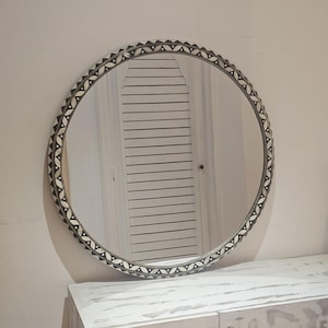 Może przedstawiać: Okrągłe lustro z ozdobną ramą z powtarzającym się wzorem czarno-białych kształtów. Lustro jest zamontowane na białej ścianie nad białą szafką z dwiema drzwiami.