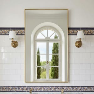 Peut inclure: Un miroir rectangulaire à cadre doré est suspendu au-dessus d'un comptoir en marbre avec deux éviers et des robinets dorés. Le miroir reflète une fenêtre avec vue sur de grands arbres verts. Deux appliques avec abat-jours blancs encadrent le miroir.