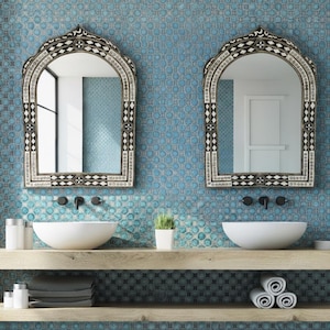 Peut inclure: Salle de bain avec deux miroirs arqués aux cadres décoratifs au-dessus de lavabos blancs. Les miroirs sont fixés sur un mur bleu à motifs. Une étagère en bois supporte les lavabos, les articles de toilette et une petite plante. Le design mélange moderne et traditionnel.