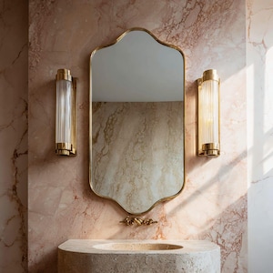 Puede incluir: Un espejo dorado con marco festoneado cuelga sobre un lavabo de piedra. Dos apliques dorados con vidrio acanalado están montados en la pared de mármol rosa. Los accesorios del baño crean una estética lujosa y vintage.