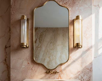 Puede incluir: Un espejo dorado con marco festoneado cuelga sobre un lavabo de piedra. Dos apliques dorados con vidrio acanalado están montados en la pared de mármol rosa. Los accesorios del baño crean una estética lujosa y vintage.