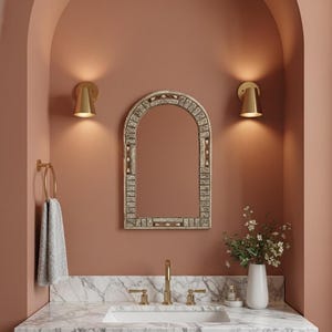 Puede incluir: Un tocador de baño con encimera de mármol, un lavabo blanco y un grifo dorado. Encima del lavabo hay un espejo decorativo con un marco blanco y beige. Dos apliques dorados flanquean el espejo y una toalla cuelga de un toallero dorado.