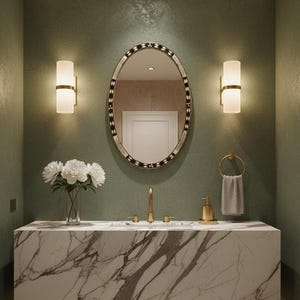 May include: A bathroom vanity with a marble countertop, gold fixtures, and an oval mirror with a decorative border. Two sconces flank the mirror, and a vase of white flowers sits on the counter. A gold towel ring is on the right.