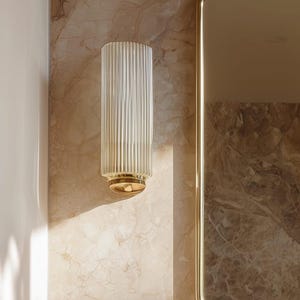 Peut inclure: Applique murale en verre transparent et cannel&eacute;, avec une base dor&eacute;e, fix&eacute;e sur un mur &agrave; motifs marbr&eacute;s. Un miroir dor&eacute; est &agrave; droite. La forme cylindrique et les rainures verticales cr&eacute;ent un effet textur&eacute;.