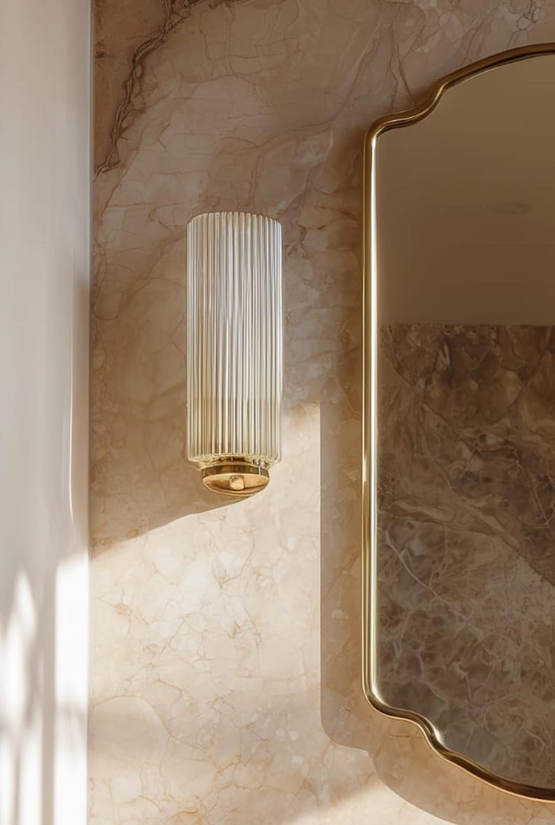 Peut inclure: Applique murale en verre transparent et cannel&eacute;, avec une base dor&eacute;e, fix&eacute;e sur un mur &agrave; motifs marbr&eacute;s. Un miroir dor&eacute; est &agrave; droite. La forme cylindrique et les rainures verticales cr&eacute;ent un effet textur&eacute;.