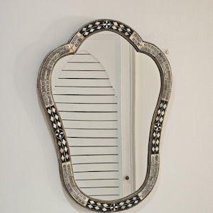 Peut inclure: Miroir décoratif orné, de forme festonnée unique. Le cadre présente un motif détaillé de formes géométriques noires et blanches, avec une bordure argentée. Le miroir est accroché à un mur blanc.