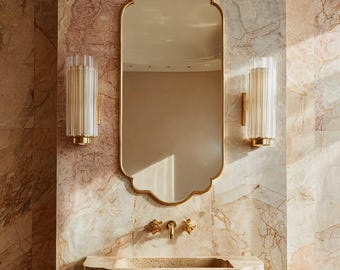 Puede incluir: Un baño con un espejo dorado con borde festoneado y apliques de pared cilíndricos. El lavabo y el grifo son dorados, sobre una pared con efecto mármol.