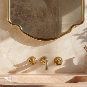 Peut inclure: Int&eacute;rieur de salle de bain avec un miroir dor&eacute; aux bords ondul&eacute;s. En dessous, des robinets dor&eacute;s, dont un robinet central et deux poign&eacute;es en forme de croix, sont fix&eacute;s sur un mur en marbre. Le lavabo est en pierre rose clair.