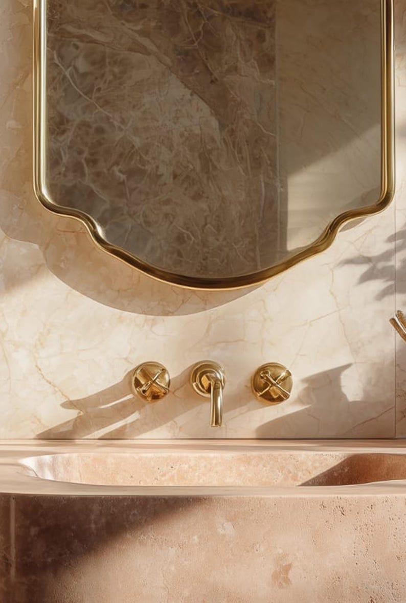 Peut inclure: Int&eacute;rieur de salle de bain avec un miroir dor&eacute; aux bords ondul&eacute;s. En dessous, des robinets dor&eacute;s, dont un robinet central et deux poign&eacute;es en forme de croix, sont fix&eacute;s sur un mur en marbre. Le lavabo est en pierre rose clair.