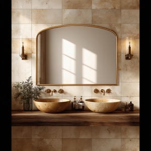 May include: A bathroom vanity featuring a large arched mirror with a gold frame, two round stone vessel sinks, and wall-mounted gold taps. The setup includes two glass wall sconces and a wooden countertop against beige tiled walls.