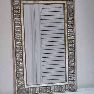 Peut inclure: Miroir rectangulaire avec un cadre décoratif. Le cadre présente un fond crème avec des détails argentés ornés. Le miroir reflète une fenêtre avec des stores horizontaux. Le design est élégant et convient à la décoration intérieure.