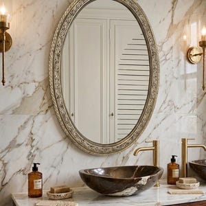 May include: An oval mirror with a decorative frame hangs above a marble countertop with a brown vessel sink. Gold faucets and sconces flank the mirror. Soap dispensers, soap, and combs complete the bathroom vanity.