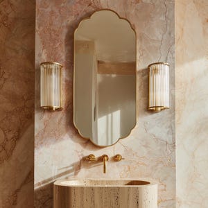 May include: A bathroom interior featuring a gold-framed, scalloped-edge mirror, flanked by two gold sconces with vertical glass rods. Below, a gold faucet and a stone sink are visible against a pink and brown marble wall.