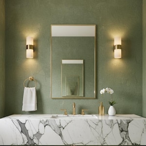 Könnte beinhalten: Ein modernes Badezimmer mit Marmor-Waschtisch und goldenen Armaturen. Über dem Waschbecken hängt ein großer rechteckiger Spiegel, der kleinere Spiegel reflektiert. Zwei Wandleuchten flankieren den Spiegel, und ein weißes Handtuch hängt an einem goldenen Handtuchring.
