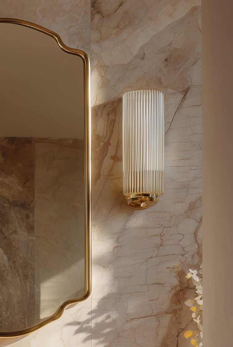 Peut inclure: Applique murale en verre transparent et cannel&eacute;, avec une base et des accents dor&eacute;s. L'applique est fix&eacute;e sur un mur &agrave; motifs de marbre, &agrave; c&ocirc;t&eacute; d'un miroir dor&eacute;. Le luminaire est de forme cylindrique.