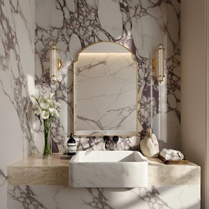 以下が含まれることがあります： 大理石のカウンタートップと長方形の洗面台を備えた豪華なバスルーム。洗面台の上には金色のフレームの鏡があり、両側に2つの壁取り付け用燭台があります。壁は紫色の縞模様の大理石で覆われています。白い花の花瓶が置かれています。