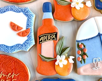 May include: Assortment of decorated sugar cookies featuring an orange and white floral design, a blue and white umbrella, and bottles of Aperol. The cookies are decorated with intricate details and bright colors.