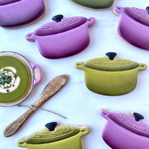 May include: A collection of six decorated sugar cookies shaped like Le Creuset Dutch ovens. The cookies are in two colours: purple and green. The cookies are arranged on a white surface.