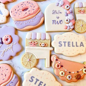 Puede incluir: Un conjunto de galletas de azúcar decoradas con temática de donuts. Las galletas están decoradas con glaseado rosa, morado y blanco y chispas. Las galletas presentan varios diseños, incluyendo donuts, un carro de donuts y las palabras "Stella tiene dos años" y "Stella".