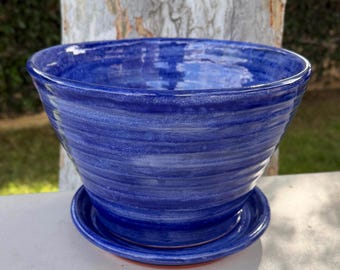 May include: A deep blue ceramic plant pot with a matching saucer. The pot features a wide, flared rim and a series of horizontal, concentric ridges along its exterior surface, finished with a glossy, variegated glaze that shows lighter blue tones.