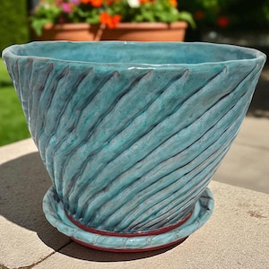 May include: A turquoise ceramic planter with a textured, spiral design. The planter has a wide, open top and sits on a matching saucer with a red rim. The outdoor setting includes other planters with flowers.