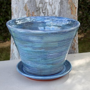May include: A wide, tapered ceramic plant pot with a matching circular saucer. The exterior features a glossy, variegated glaze in shades of deep navy, sky blue, and turquoise, with horizontal ridged textures circling the entire body of the vessel.