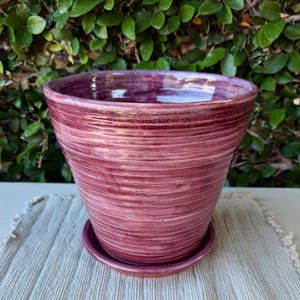 May include: A deep magenta ceramic plant pot with a wide, flared rim and a matching circular saucer. The exterior surface features a series of horizontal, grooved ridges that wrap around the body, creating a textured, layered appearance.