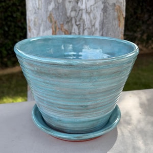 May include: A turquoise ceramic planter with a matching saucer. The planter has a wide, flared shape with a textured, swirled design. The saucer has a red rim. The planter is suitable for indoor or outdoor use.