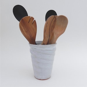 May include: A white ceramic utensil holder with a textured, swirled design. It contains wooden cooking spoons and a black spatula. The spoons are various shapes and sizes, and the spatula is a solid black color.
