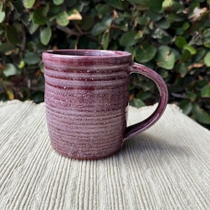 May include: A cylindrical ceramic mug featuring a deep burgundy glaze with a speckled, textured finish. The exterior surface displays horizontal ridged detailing throughout, and the mug includes a single curved handle on the right side.