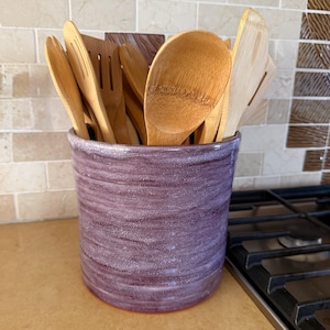 May include: A cylindrical ceramic utensil holder with a textured, speckled purple glaze finish. The container is filled with a variety of light brown bamboo kitchen utensils, including several wooden spoons and a slotted spatula, standing upright.