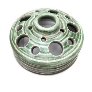 May include: A green ceramic flower frog with multiple holes for arranging flowers. The frog has a textured, glazed surface and a circular base.
