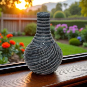 May include: A decorative vase with a textured, swirling design. The vase is a dark gray color with horizontal ridges around the neck. The vase sits on a wooden surface with a blurred outdoor background.