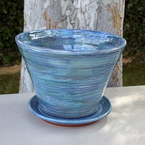 May include: A handcrafted ceramic planter in shades of blue and teal, with a matching saucer. The planter has a wide, flared shape and a textured surface. The saucer has a red rim. Ideal for indoor or outdoor use.
