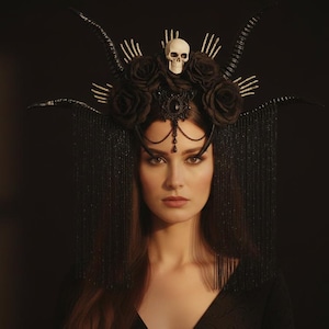 May include: A woman wearing an elaborate black headpiece featuring a skull, roses, and beaded fringe. The headpiece is the central element, set against a dark backdrop. The woman gazes directly at the camera.
