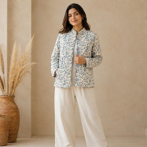 Könnte beinhalten: Eine gesteppte Jacke mit blau-weißem Blumenmuster, Stehkragen, langen Ärmeln und Vordertaschen, getragen über einem schlichten weißen Oberteil und einer cremefarbenen weiten Hose mit braunen offenen Sandalen.